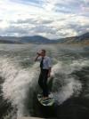 guy in formal clothing surfs and drinks a beer behind a boat in a lake under beautiful mountains