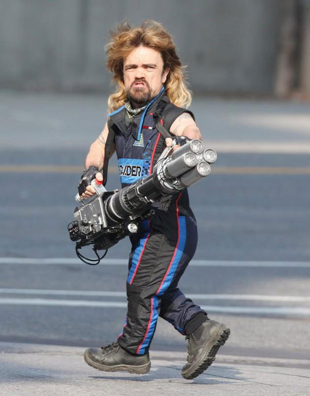 peter dinklage with a mullet wearing a sleeveless jumpsuit and holding a four-barrel laser cannon
