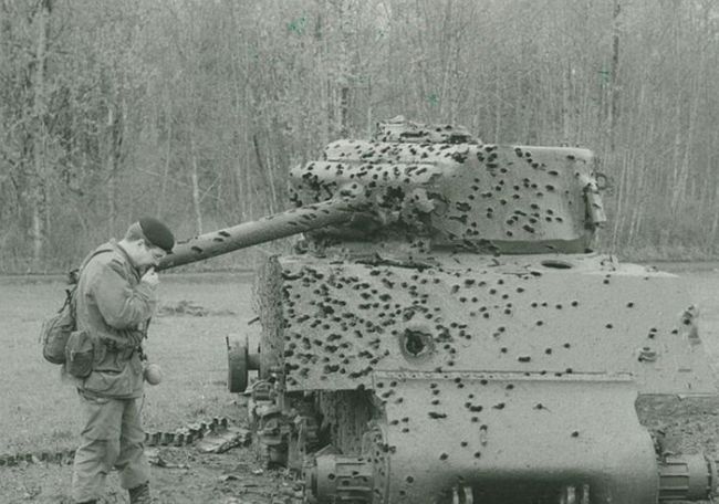 black and white photograph of a tank riddled with bullet holes and a soldier standing next to it