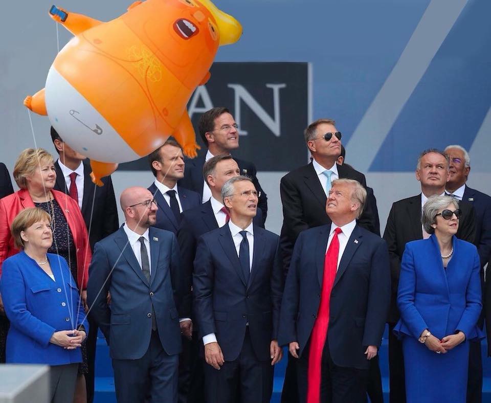 trump looking at baby trump balloon held by merkel